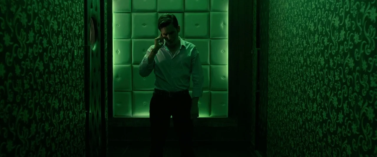 The Raid 2 (2014) movie still: fullbody — An interior space features patterned dark wallpaper and a bright green glowing panel. A man stands…
