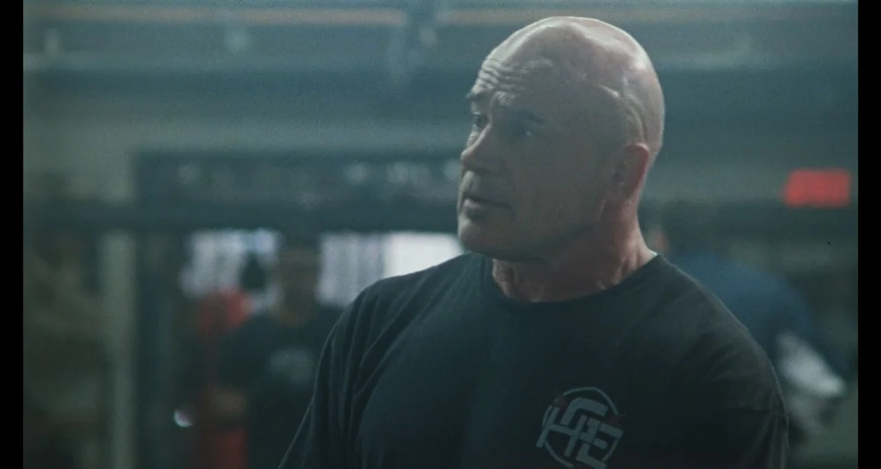 The Smashing Machine (2025) movie still: medium — A dimly lit workout facility features blurred exercise equipment and metal structures in…