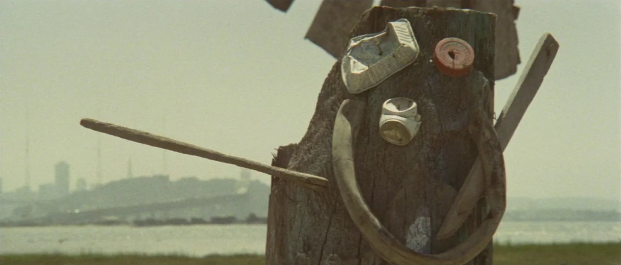 Take Me Away! (1978) movie still: closeup — A weathered wooden post stands near a coastline with a city visible in distance. Scraps of trash…