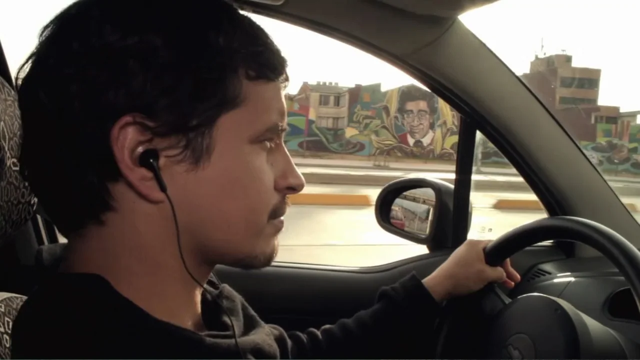 Aquel Septiembre (2018) movie still: medium — The interior of a moving car shows a driver looking out the window. A colorful mural on a building…