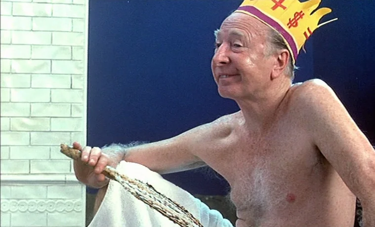 The Garden (1990) movie still: medium — An elderly man sits in a room with white brick walls and blue panels. He is wearing a paper crown and holds…