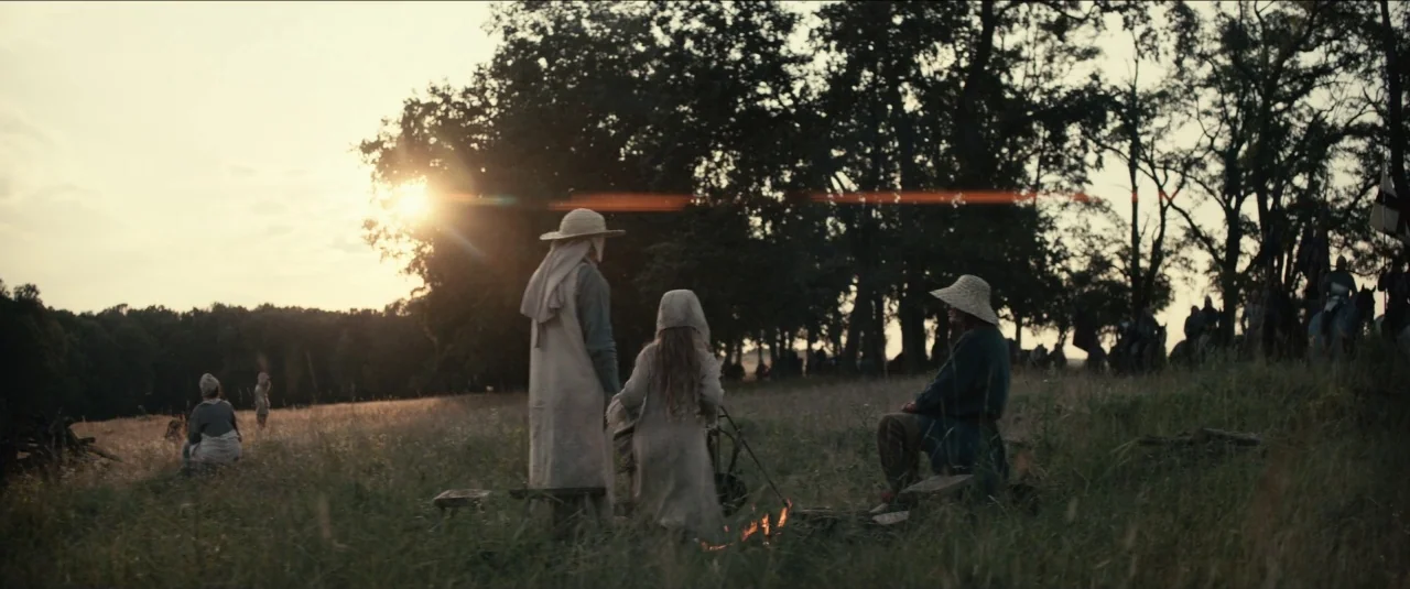 The King movie still: wide — A grassy field with a line of trees sits under a sunset sky. Three people stand and sit around a small fire as others…