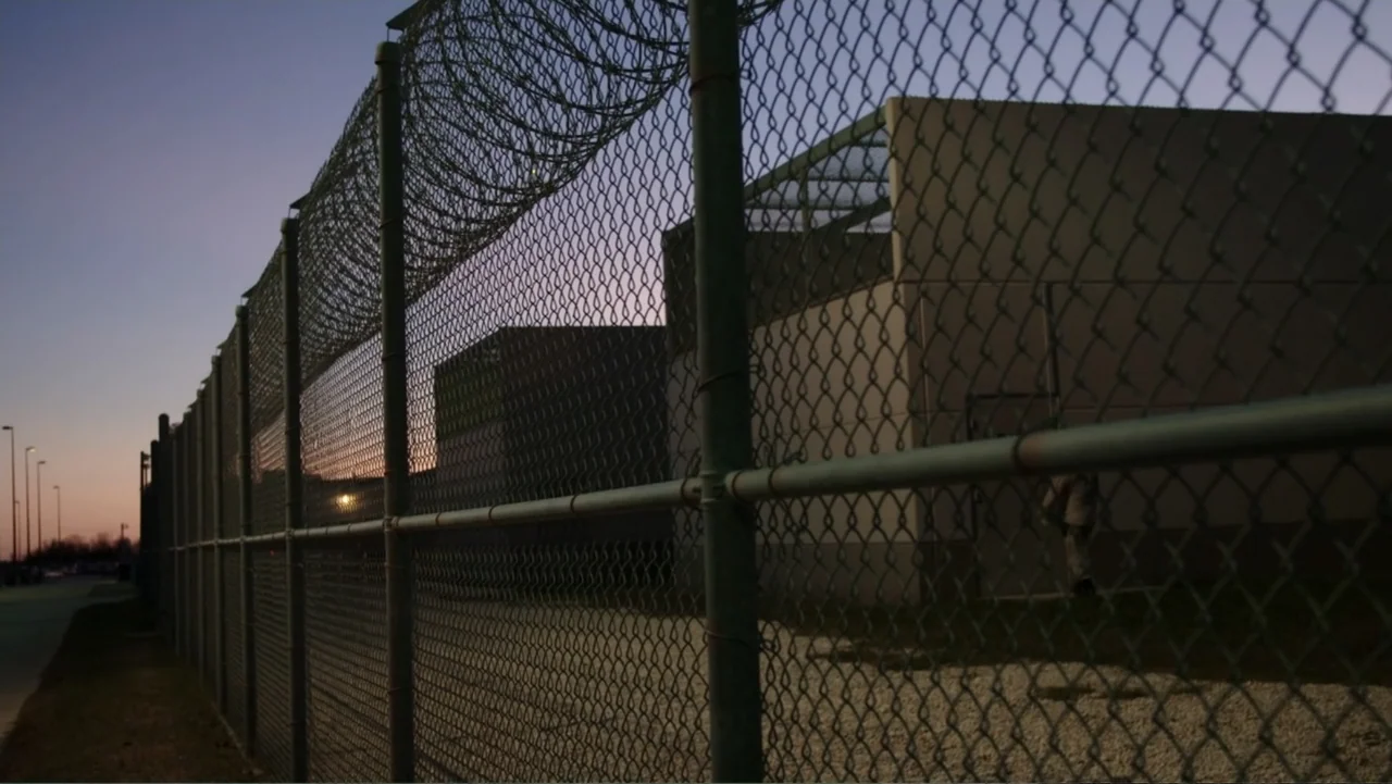 Torn Apart: Separated at the Border (2019) movie still: wide — A tall chain-link fence topped with barbed wire encloses a concrete yard at dusk. The…