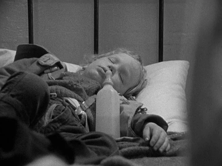 Cathy Come Home (1969) movie still: medium — A child sleeps in a simple iron-framed bed with white pillows and blankets. A glass baby bottle rests…