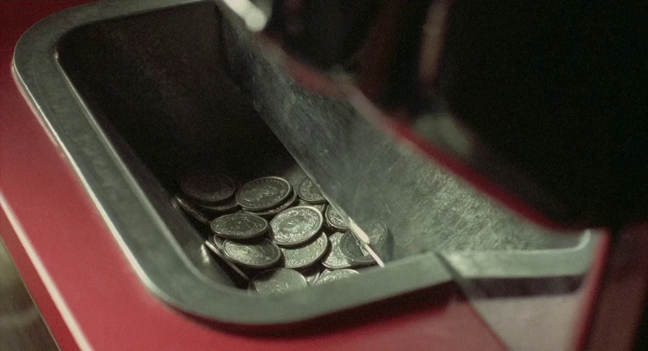 Three Colours: Red (1994) movie still: closeup — A pile of silver coins sits inside the steel retrieval bin of a machine. The surrounding casing is…