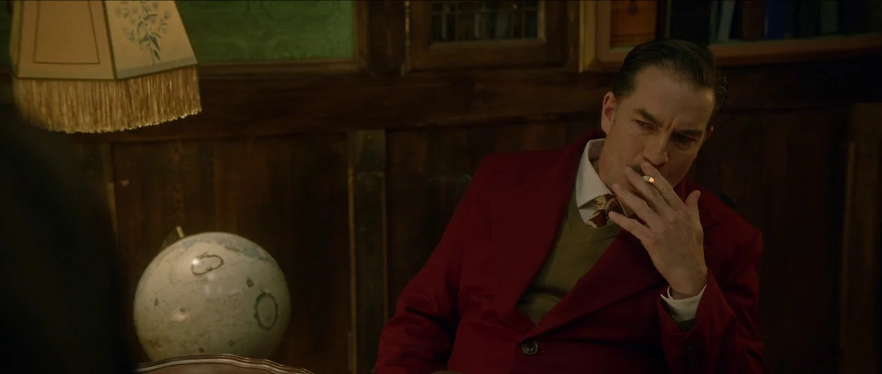 Brooklyn 45 (2023) movie still: medium — A dark study features wooden paneled walls, a fringed lampshade, and a spherical globe. A man in a red…
