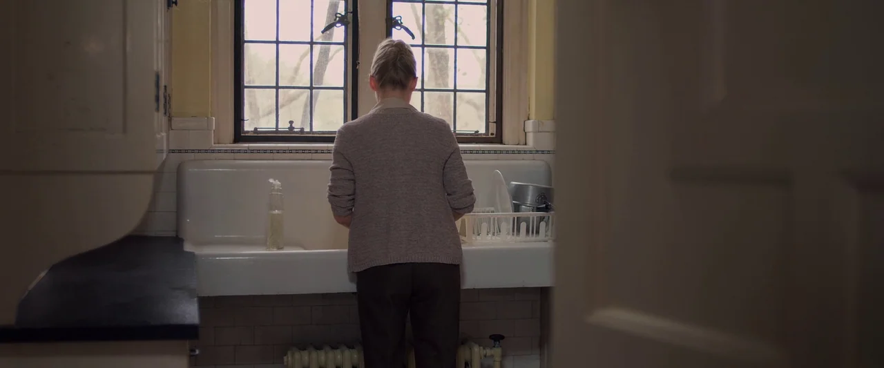 You’re Next (2013) movie still: medium — A kitchen features a white porcelain farmhouse sink positioned below a divided-light window. A woman stands…