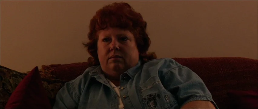 Bubble (2005) movie still: medium — A dimly lit interior space with a person seated on a sofa. The individual gazes forward with a neutral…