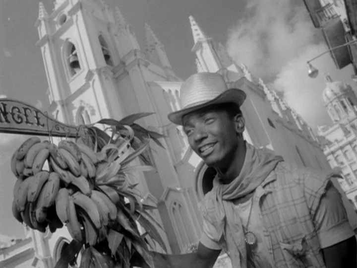 I Am Cuba (1964) movie still: medium — A man stands on a sidewalk in front of an ornate cathedral with white spires. He holds a long pole draped…