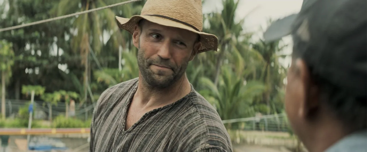 The Meg (2018) movie still: medium — An outdoor location with blurred green foliage and palm trees in the background. A man wearing a straw hat…