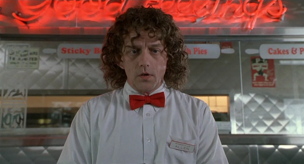 Ghost World (2001) movie still: medium — A classic 1980s diner setting with red neon signage visible above the counter area. A male employee in a…
