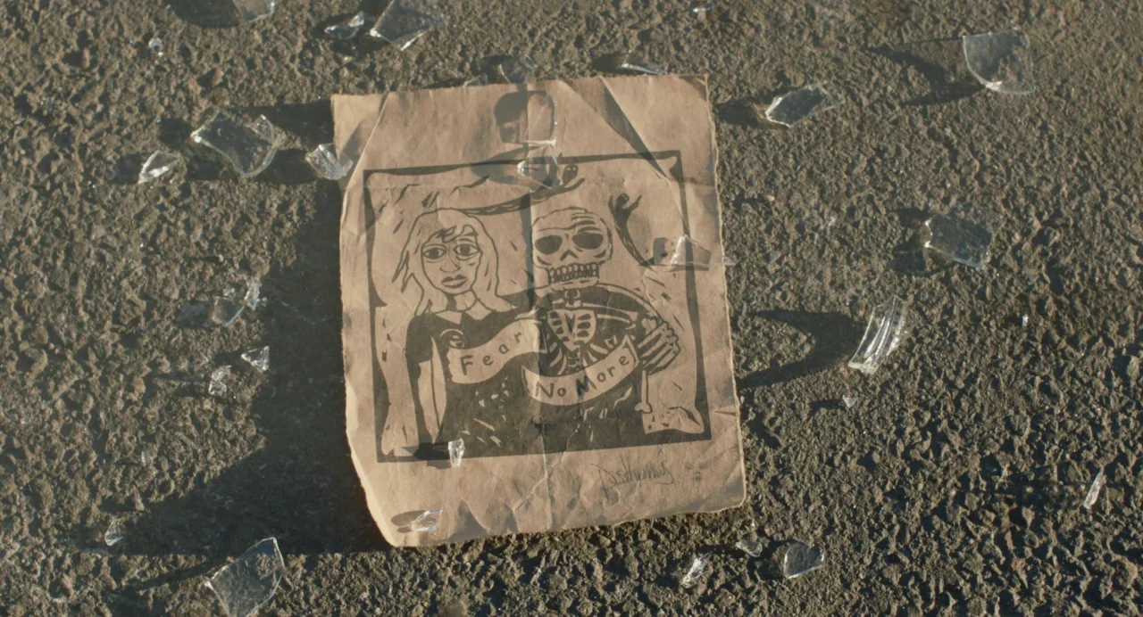 Cymbeline (2014) movie still: closeup — A crumpled piece of brown paper lies flat on a rough gray asphalt surface. Shards of broken clear glass are…