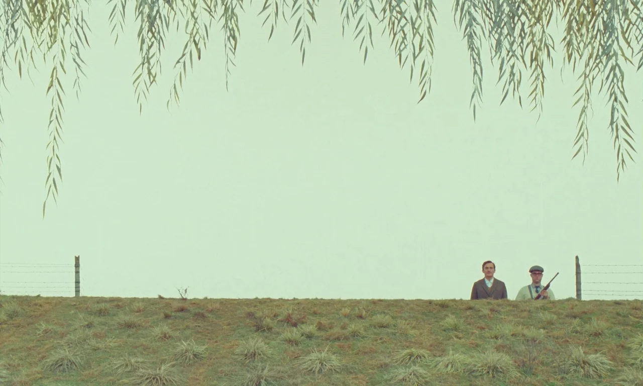 The Swan (2023) movie still: wide — A grassy embankment sits under a pale sky framed by overhanging willow branches. Two men stand in the…