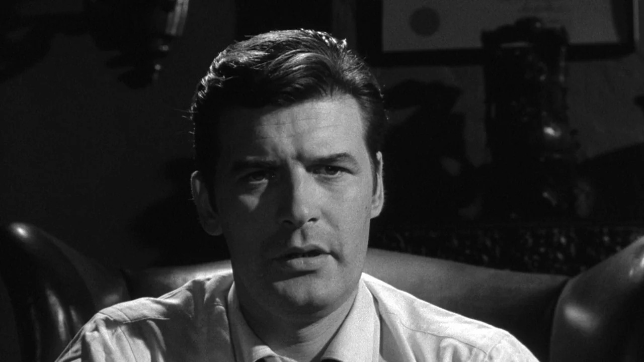 Shock Corridor (1963) movie still: closeup — A dark office setting with framed certificates visible on the wall in the background. A man sits in a…