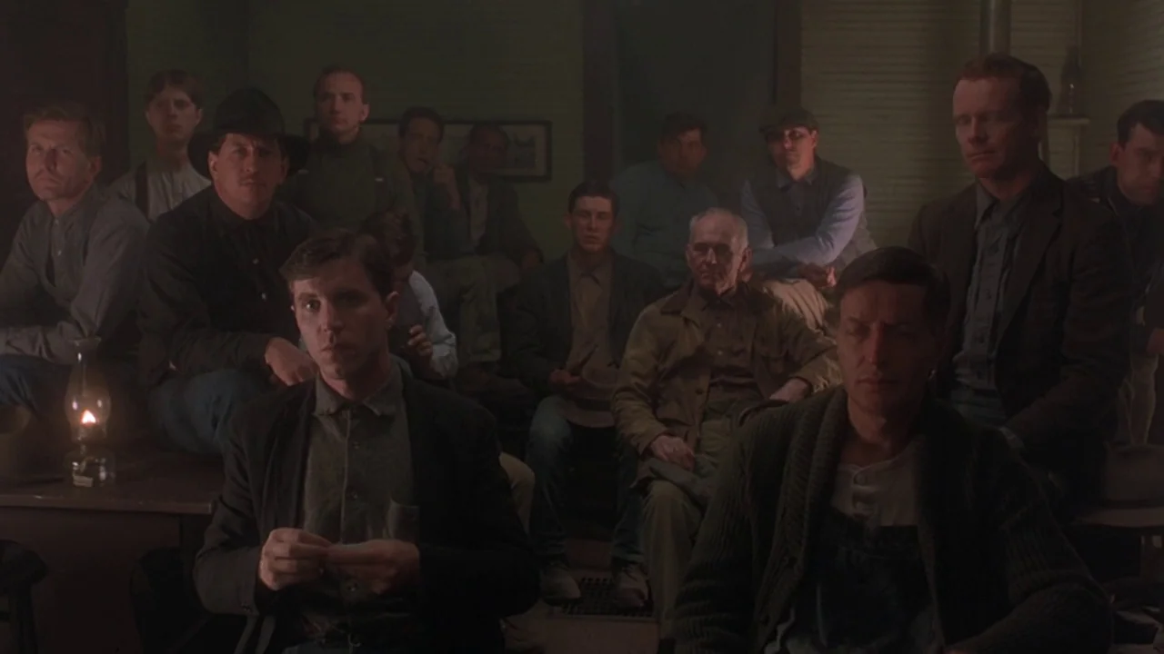 Matewan (1987) movie still: medium — A dimly lit room filled with men sitting in rows on wooden chairs and benches. The men watch an unseen speaker…