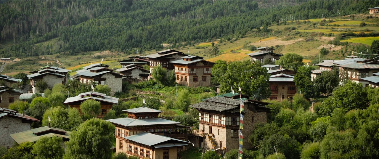 The Monk and The Gun (2023) movie still: establishing — A cluster of traditional Bhutanese stone houses sits nestled in a mountainous green valley.…