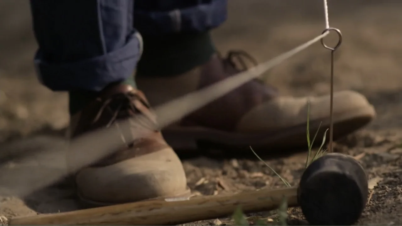 Like.Share.Follow. (2017) movie still: closeup — Dry dirt ground features a metal stake with a vibrating string attached to it. A mallet lies nearby…