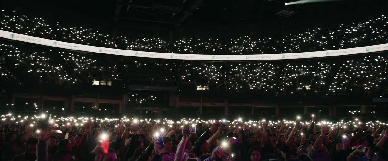 Making of: F*CKS NEWS (2025) movie still: wide — A dark indoor arena is filled with a large crowd in the lower foreground. Hundreds of people hold…