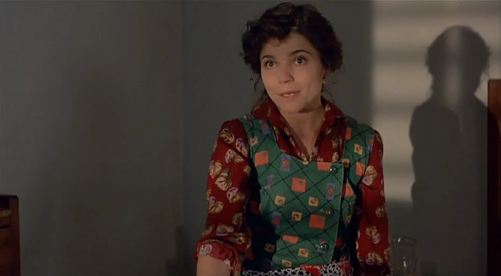 The Voice of the Moon (1990) movie still: medium — A dimly lit room features a woman standing against a plain, dark-painted wall. She faces slightly…