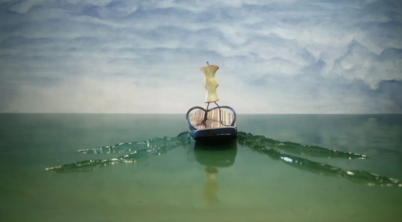 Eagle Vs. Shark movie still: medium — A miniature apple core figure stands on a floating flip-flop in calm water. The backdrop consists of a painted…