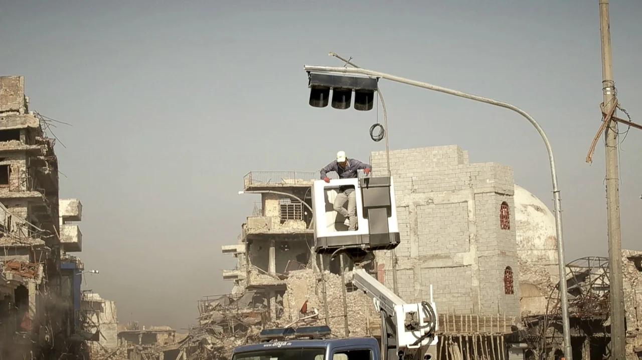 City of Two Springs (2019) movie still: medium — A worker in a cherry picker repairs a traffic light in a war-torn urban area. Damaged concrete…