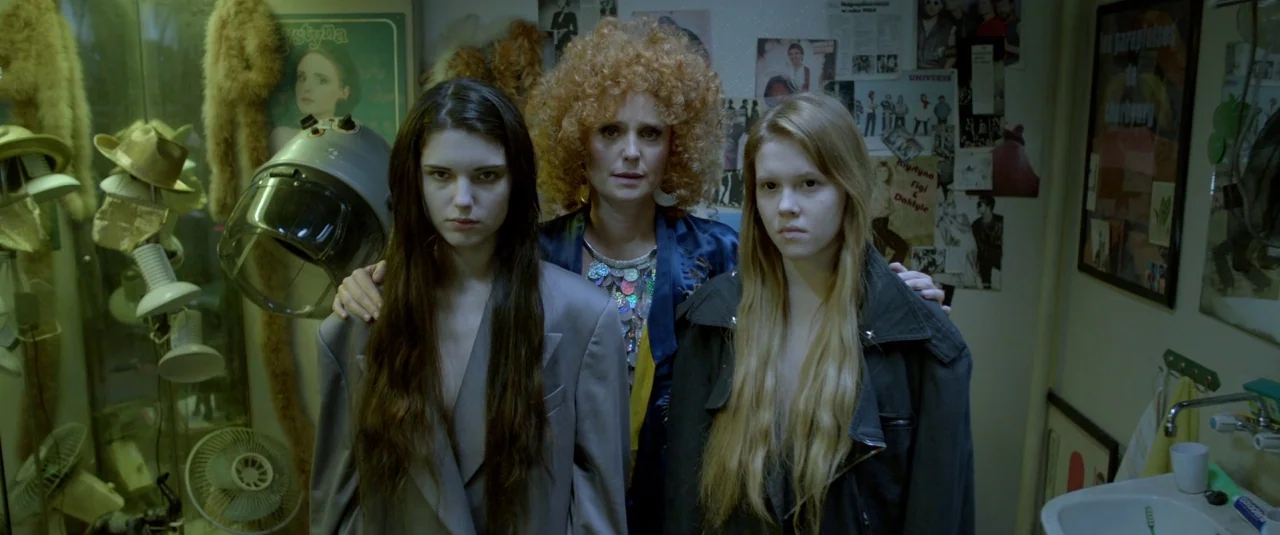 The Lure (2015) movie still: medium — A cluttered hair salon interior filled with vintage styling equipment and wall posters. Three women stand…