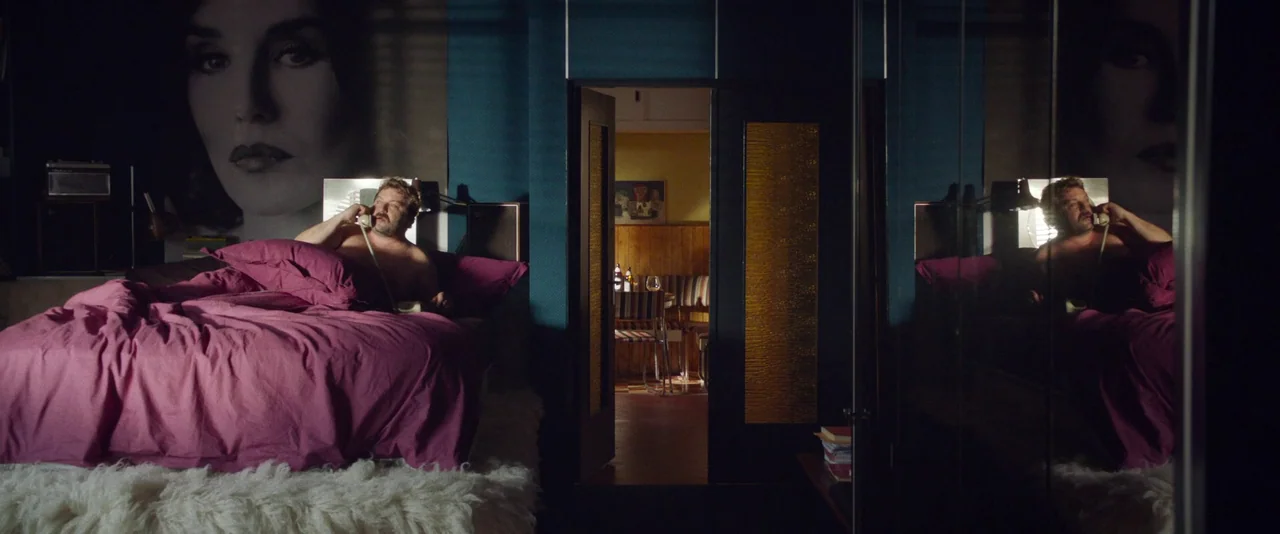 Peter Von Kant (2022) movie still: wide — A dark, blue-walled hotel room features a large magenta-covered bed and a vanity mirror. A man sits…