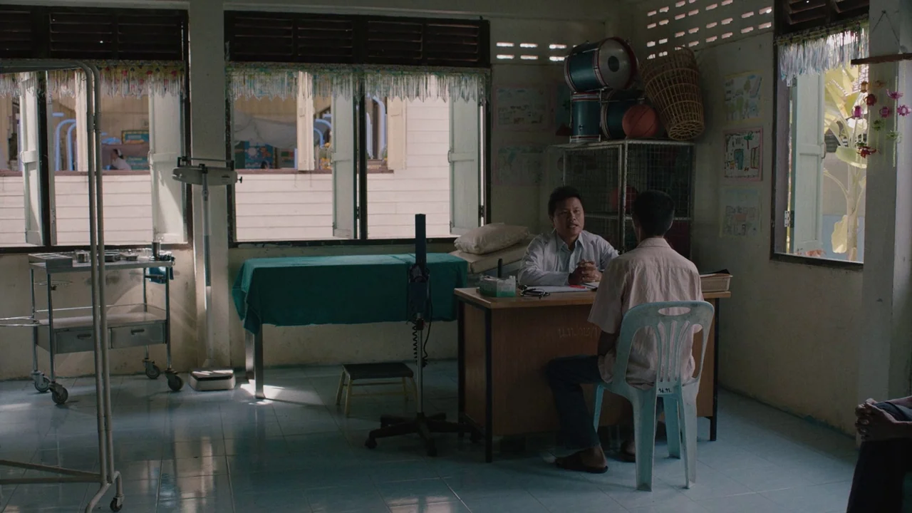 Cemetery of Splendour (2015) movie still: wide — A sparse medical office with tiled floors and windows covered by thin patterned curtains. A man…