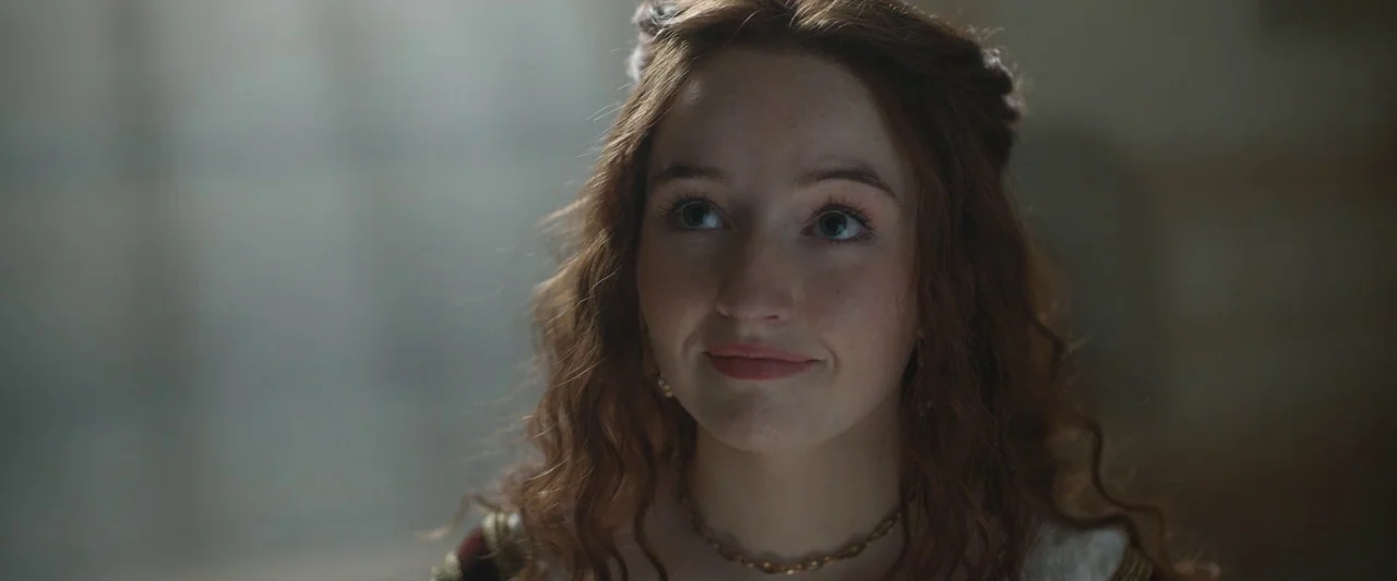 Rosaline (2022) movie still: closeup — A stone interior lit by diffuse light from a nearby window. A woman with wavy auburn hair looks slightly…