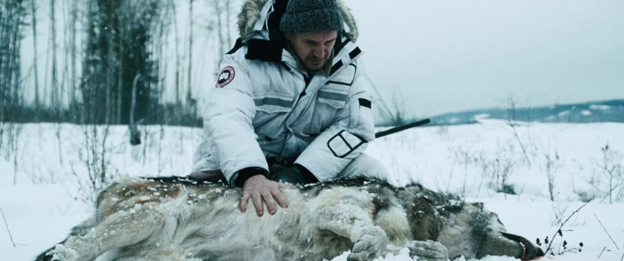 The Grey (2012) movie still: medium — A desolate, snowy landscape features sparse, thin trees in the background under an overcast sky. A middle-aged…