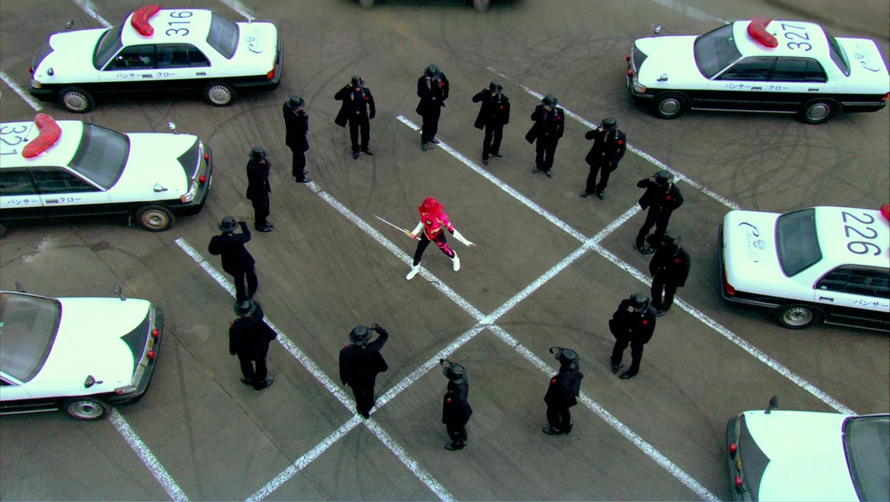 Cutie Honey (2004) movie still: wide — A paved lot marked with white lines holds several black and white patrol cars. A lone figure in a vibrant red…