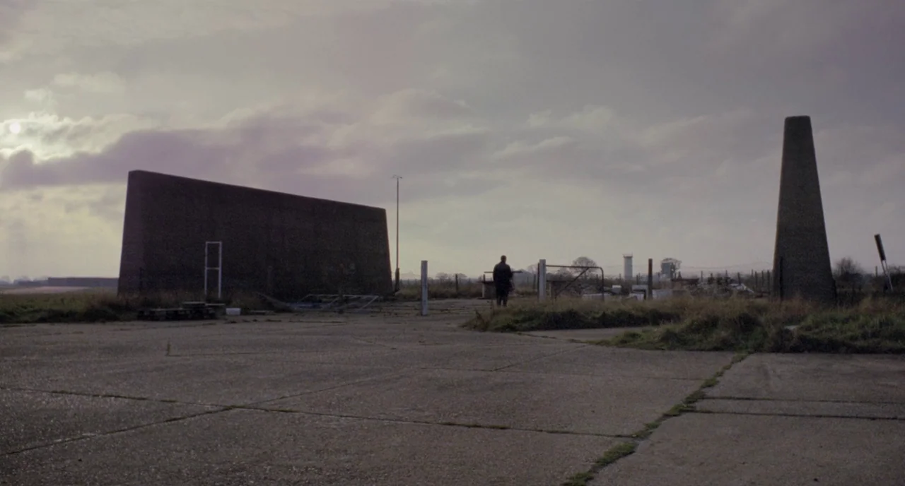 Possum (2018) movie still: wide — A vast, abandoned concrete airfield features a large, slab-like structure and a conical monolith. A lone figure…
