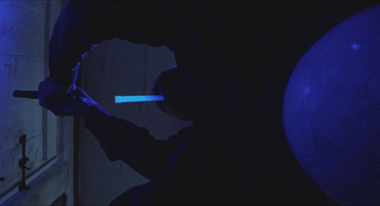 Dog Soldiers (2002) movie still: medium — A dark, ambiguous interior space features a person working in deep shadow. They bite a bright blue glow…