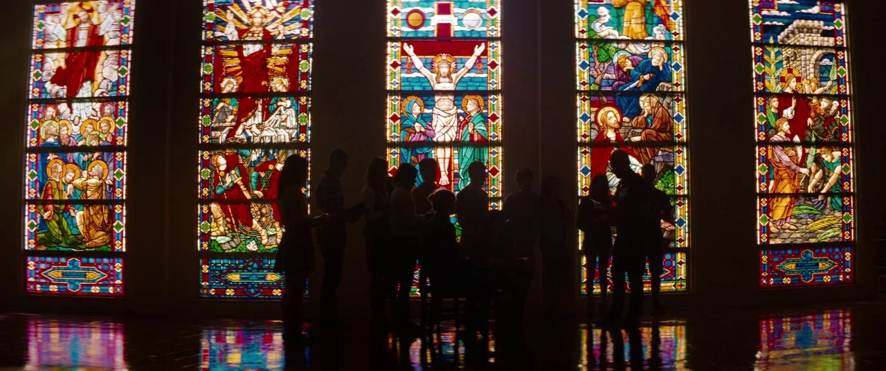 Spring Breakers (2012) movie still: wide — A group of people stand in a dark interior space with stained glass. The tall windows display colorful…