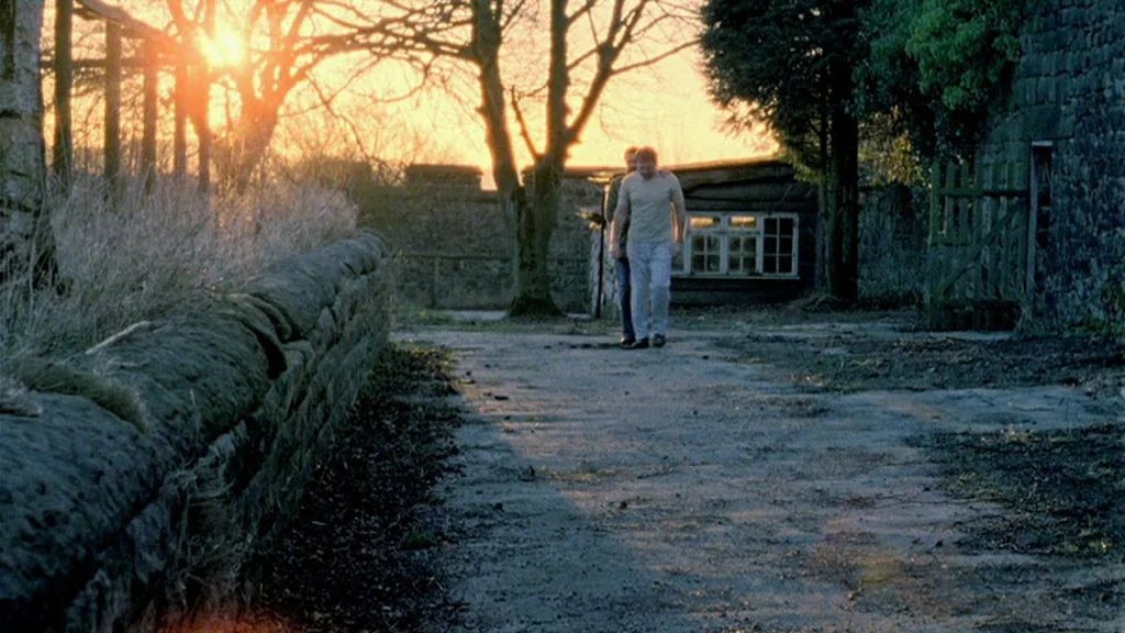 Dead Man’s Shoes (2004) movie still: wide — A rural dirt path sits between a dry stone wall and a wooden structure. Two men walk together towards…