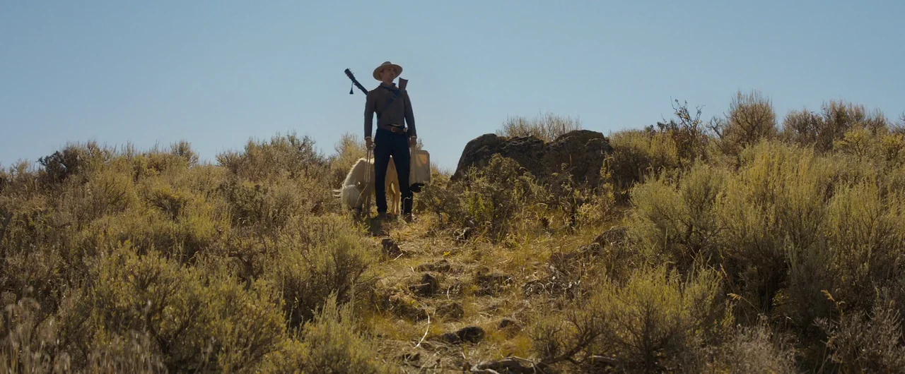 Damsel (2018) movie still: fullbody — A man stands in a dry, open field filled with sagebrush under a clear sky. He wears a cowboy hat and carries a…