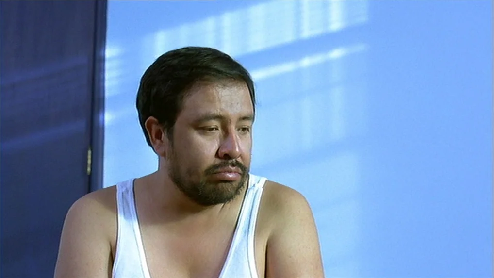Battle In Heaven (2005) movie still: medium — A man stands before a solid blue wall marked by light through window blinds. The man wears a white…