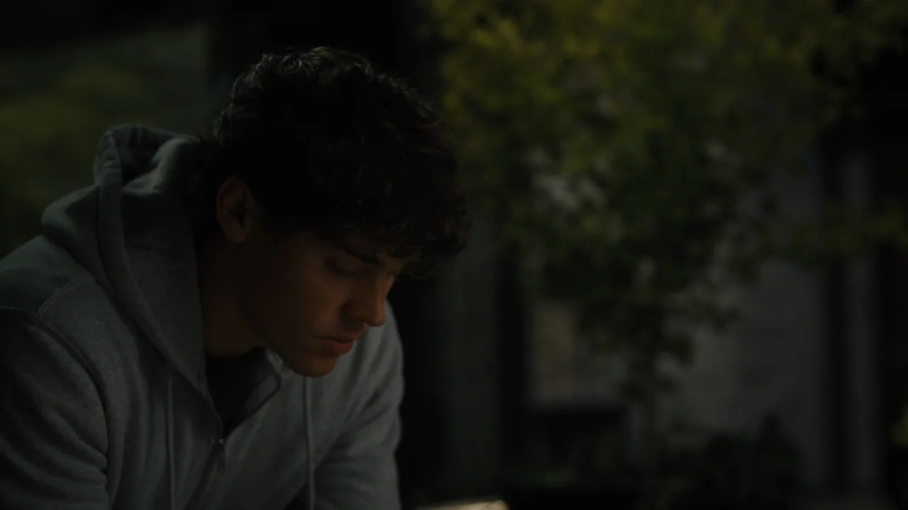 Sidelined: The QB and Me (2025) movie still: medium — A man in a hooded sweatshirt sits outdoors at night against a blurred leafy background. He is…