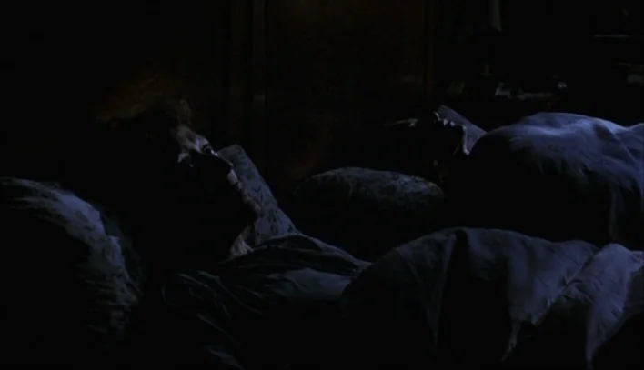The Piano Teacher (2001) movie still: medium — A dark bedroom holds a couple lying in bed beneath heavy blankets. The man stares upward while the…