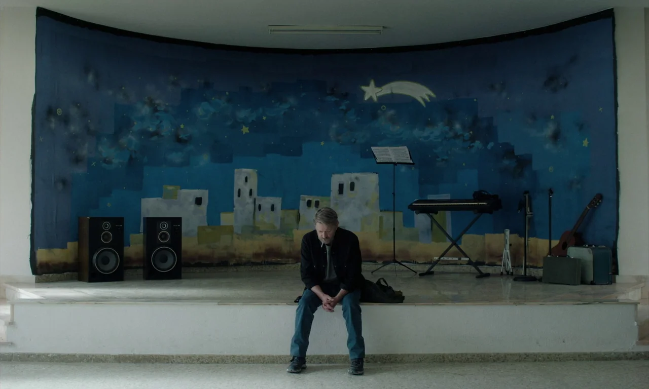 Close Your Eyes (2023) movie still: fullbody — A man sits on a low stage fronted by a hand-painted blue night sky mural. Musical equipment including…