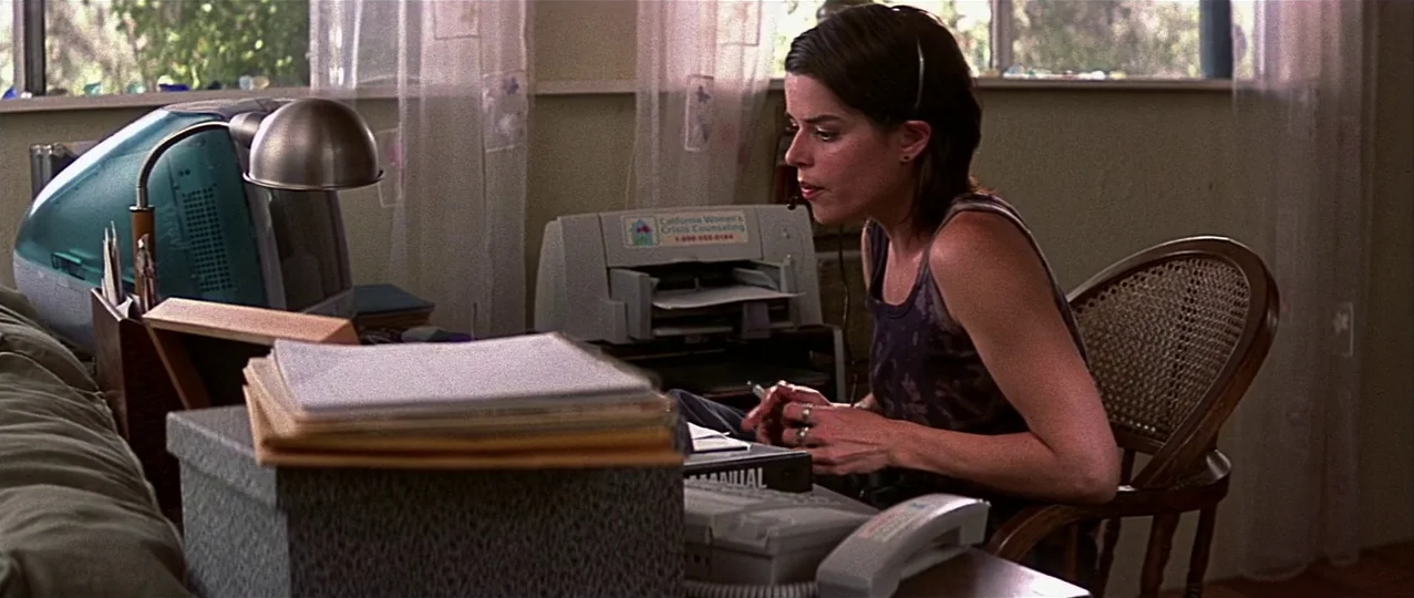 Scream 3 (2000) movie still: medium — A desk with a computer and papers sits in a home office near a window. A woman sits in a wooden chair working…