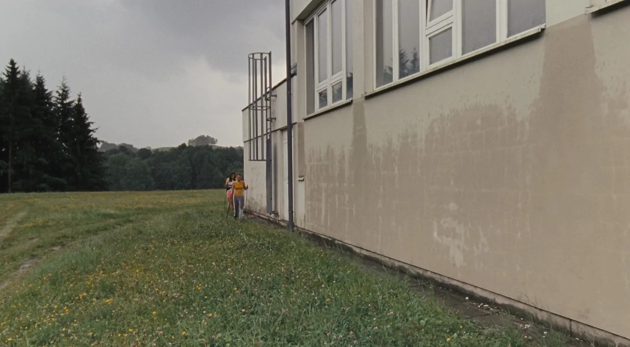 Paradise: Hope (2013) movie still: wide — A concrete building wall stands adjacent to a grassy field with a distant treeline. Two women walk forward…