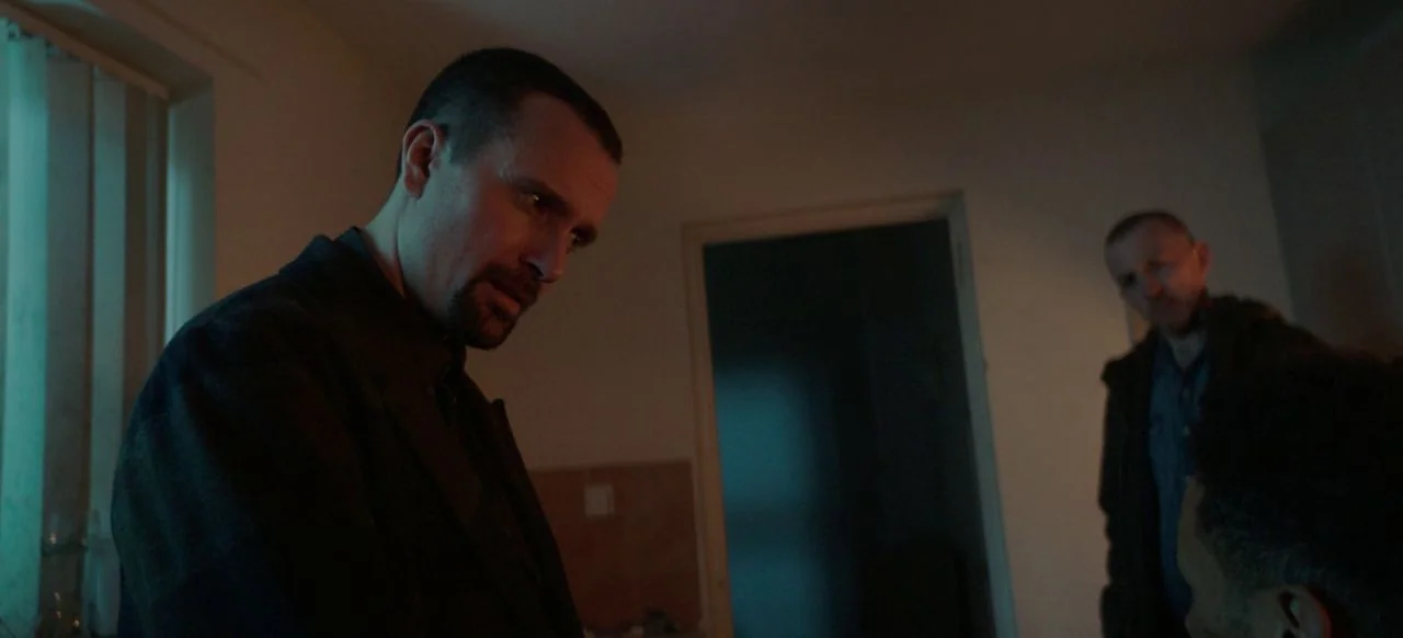 A Good Woman Is Hard To Find (2019) movie still: medium — A dimly lit room features a doorway and a window with vertical blinds. Two men occupy the…