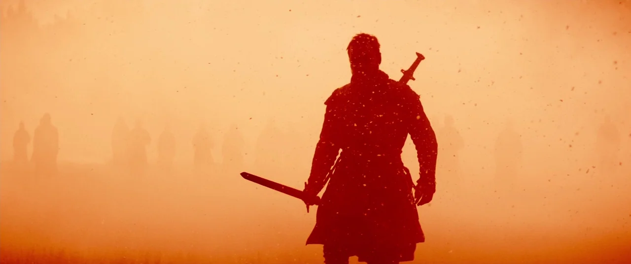 Macbeth (2015) movie still: fullbody — A field filled with orange dust and silhouettes of distant standing figures. A foreground soldier in dark…