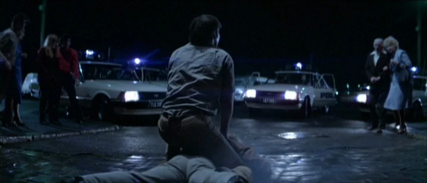 Roadgames (1981) movie still: fullbody — A nighttime scene in a parking lot with two police cars and multiple bystanders. A man kneels over another…