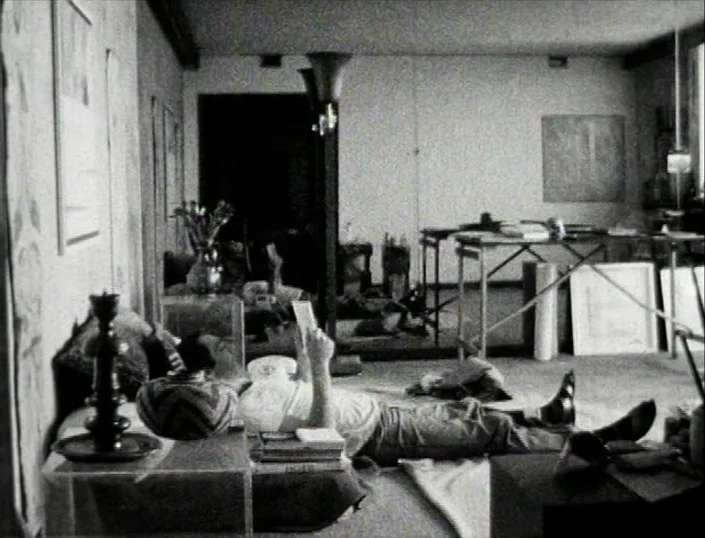 Glitterbug (1994) movie still: wide — An artist's studio contains work tables, assorted furniture, and various sketches on the walls. A person lies…