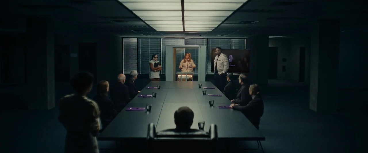 We Are Zombies (2023) movie still: wide — A dark boardroom features a long central table surrounded by seated figures. A woman sits in a…
