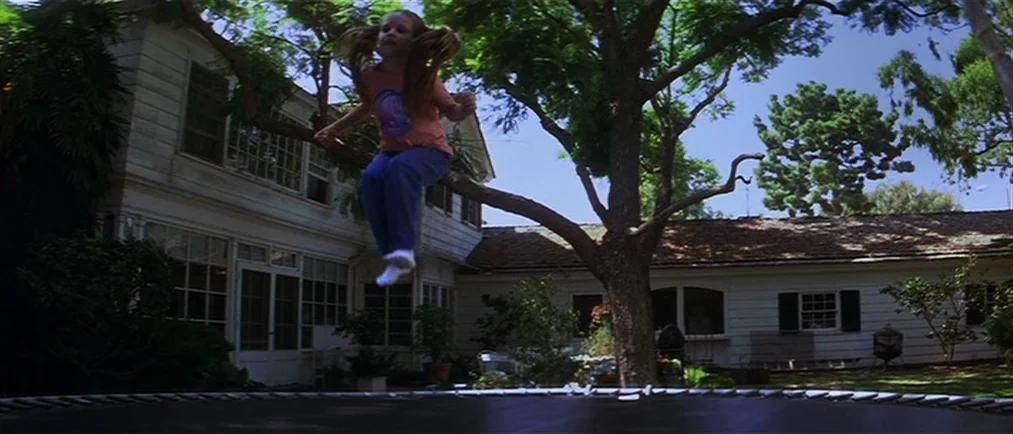 Donnie Darko movie still: fullbody — A residential white house stands in the background behind a large tree. A young child is mid-air while jumping…