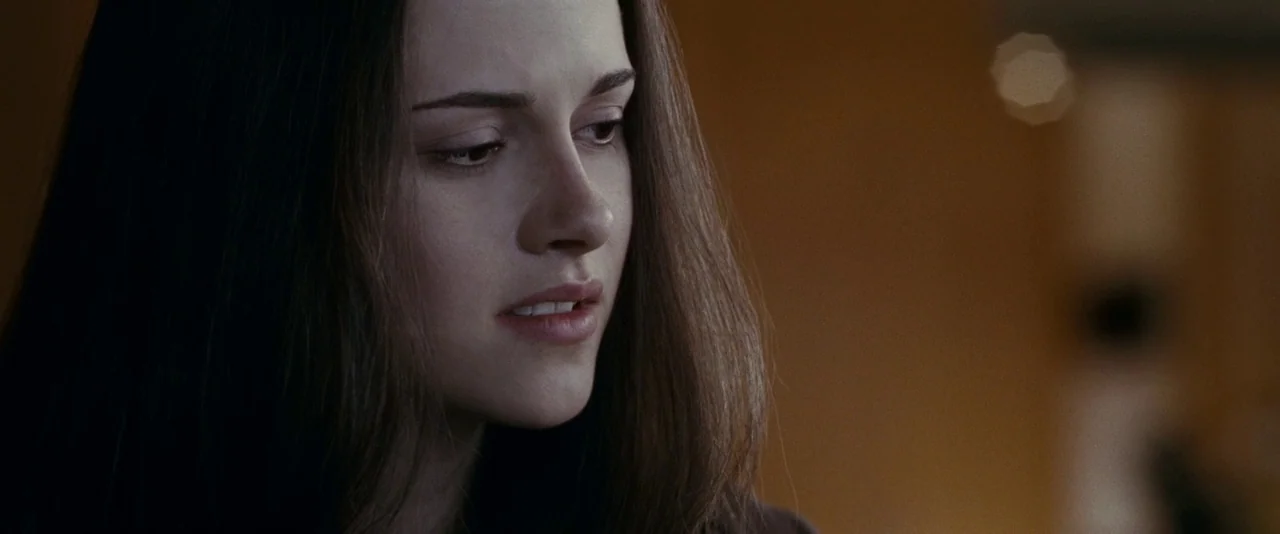 The Twilight Saga: Eclipse (2010) movie still: closeup — A woman stands in a dimly lit room with brown wall tones. She looks downward with an…