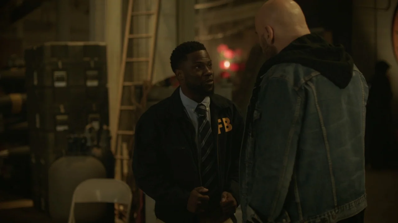 Die Hart (2023) movie still: medium — A dark, cluttered industrial space contains stacked heavy cases and a wooden ladder. An FBI agent talks to a…